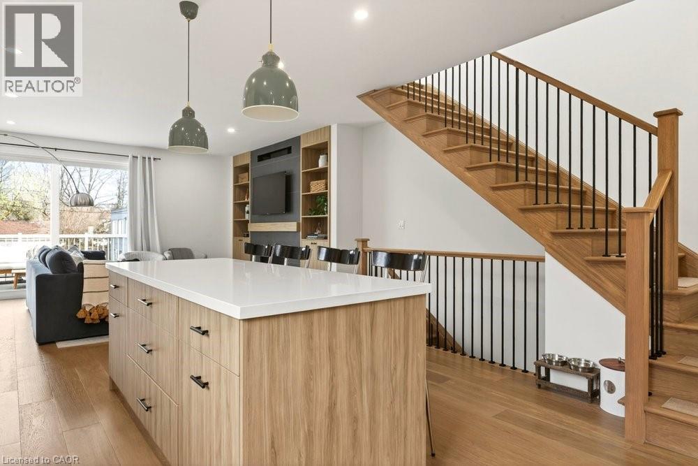 Open-concept living space featuring wood-finish flooring, a contemporary kitchen island with a light-toned countertop, a built-in media center with shelving, and a prominent staircase with wood treads