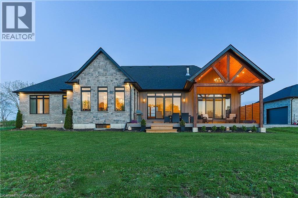 Rear exterior featuring a stone facade, a covered patio with exposed wood beams, multiple large windows, and a manicured lawn