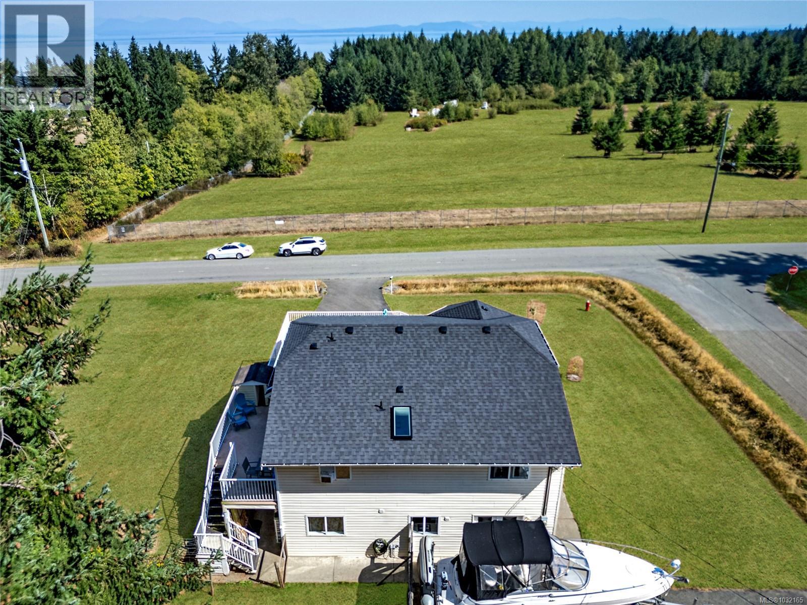 Aerial view of June Rd looking out to the Strait of Georgia and the mainland mountains beyond. The home sits a short walk from the beach, with  DND reserve land directly across the road keepi