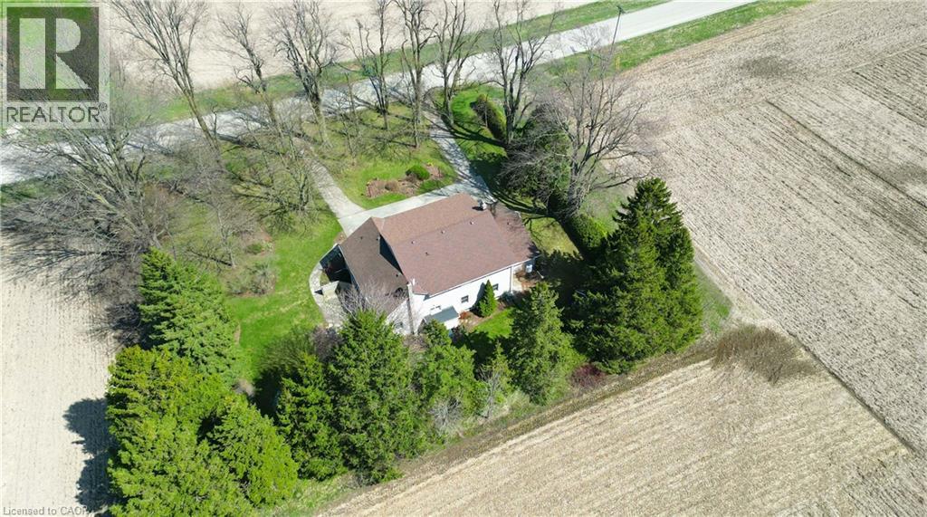 Brown shingle roof home surrounded by mature evergreen and deciduous trees, with a gravel driveway and surrounding agricultural fields