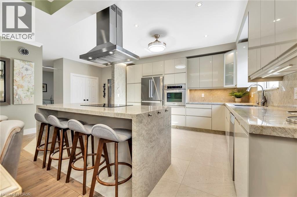 Gourmet kitchen featuring a large waterfall island with built-in cooktop, stainless steel range hood, integrated stainless steel appliances, contemporary cabinetry, and light-toned tile flooring