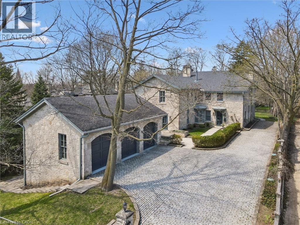 Stone residence featuring a multi-car garage, extensive paver driveway, pitched shingle roof, and mature perimeter trees