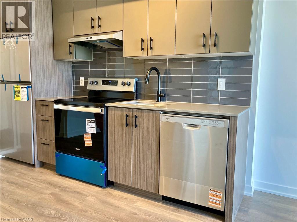 Modern kitchen featuring stainless steel appliances, grey subway tile backsplash, light-toned countertops, wood-finish flooring, and two-tone cabinetry with matte black hardware