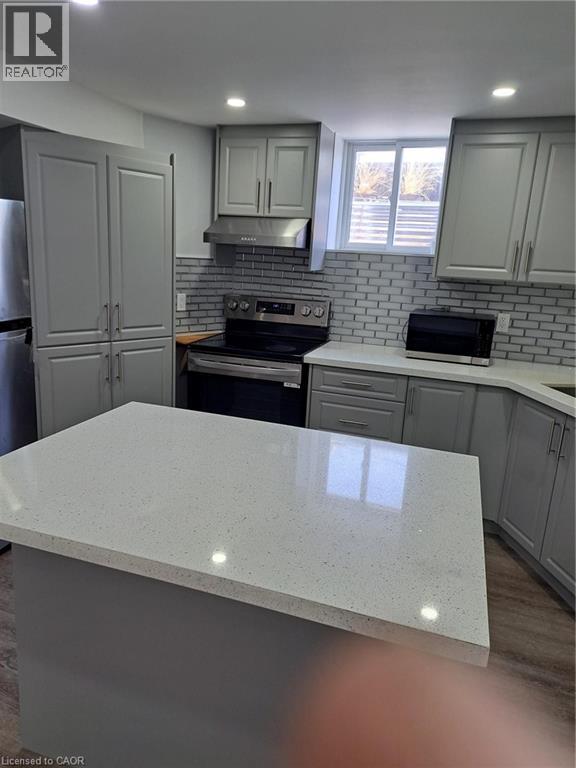 Modern kitchen featuring a spacious center island with a light-toned countertop, gray cabinetry, subway tile backsplash, stainless steel range with hood, and wood-finish flooring