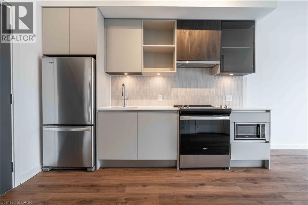 Contemporary kitchen featuring wood-finish flooring, stainless steel appliances, light-toned cabinetry, a light-colored countertop, and a tiled backsplash with under-cabinet lighting
