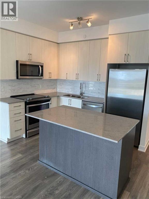 Modern kitchen featuring a central island with a light-toned countertop, stainless steel appliances, light-colored cabinetry, a tile backsplash, and wood-finish flooring