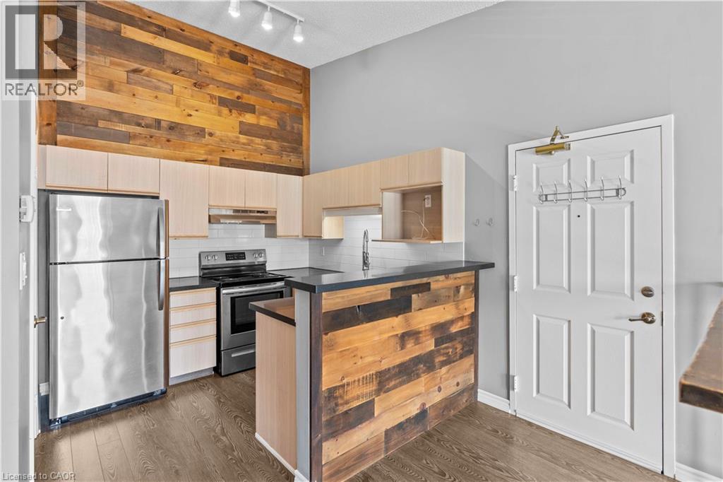 Contemporary kitchen featuring a rustic wood accent wall, stainless steel appliances, light wood-finish cabinetry, dark countertops, and wood-finish flooring