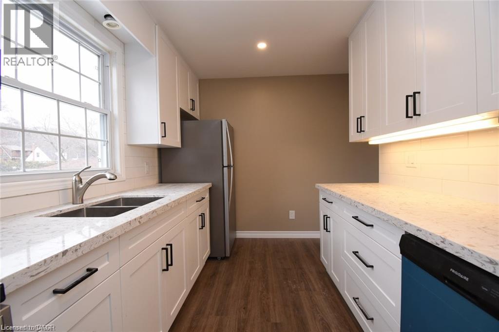 White shaker cabinetry with black hardware, stone-finish countertops, stainless steel double basin sink with gooseneck faucet, subway tile backsplash, and wood-finish flooring