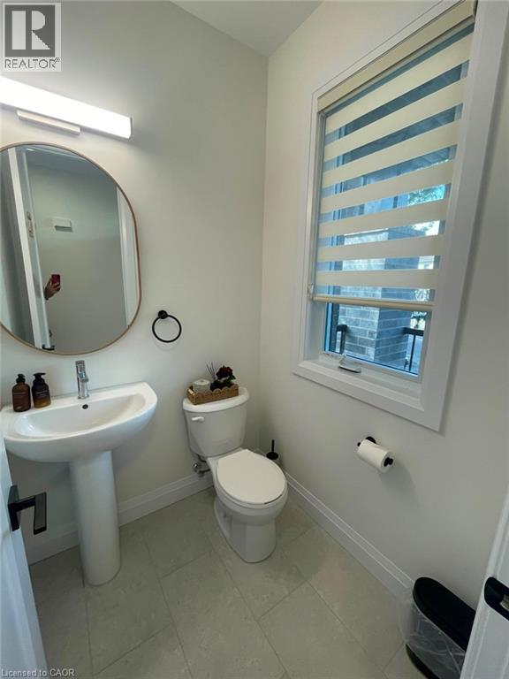 Pedestal sink, wall-mounted mirror, modern window treatment, white toilet, and light-toned floor tiling